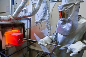 A person in a silver thermal protection suit with a protective mask and gloves uses a long metal rod to retrieve a red-hot metal box containing material samples from a hot test furnace in the laboratory of OWI Science for Fuels gGmbH.